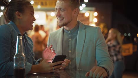 Schönes-junges-Paar-ist-mit-einem-Smartphone,-während-an-einem-Tisch-in-einem-Outdoor-Street-Food-Cafe-sitzen.-Sie-surfen-im-Internet-oder-in-sozialen-Medien,-schauen-sich-Videos-an.-Sie-sind-glücklich-und-lächeln.