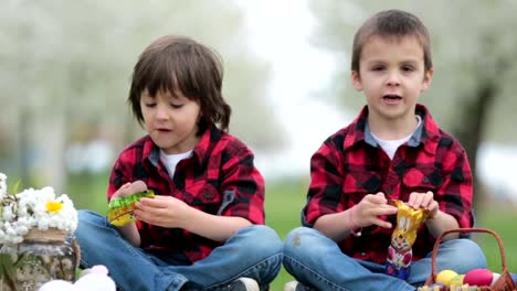 Dos-niños,-hermanos,-comiendo-conejitos-de-chocolate-y-divirtiéndose-con-huevos-de-pascua-en-el-parque,-hermoso-jardín-floreciente-primavera