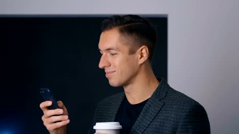 Confident-businessman-drinks-coffee-and-uses-a-smartphone-in-the-office-during-a-coffee-break