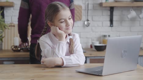 Portrait-of-cute-Caucasian-female-teenager-looking-at-laptop-screen,-holding-head-with-hand-and-smiling.-Pretty-girl-resting-at-kitchen-while-father-cooking-at-the-background.-Morning,-breakfast,-adolescence.