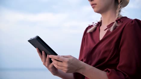 Close-up-An-attractive-young-girl-hands-in-a-summer-red-dress-sits-on-a-stone-by-the-sea-in-the-evening-and-looks-at-something-on-a-tablet.-Swipe-across-the-screen