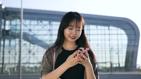 Agradable-despreocupada-joven-asiática-mujer-escribiendo-sms-en-su-teléfono-cerca-del-edificio-del-aeropuerto