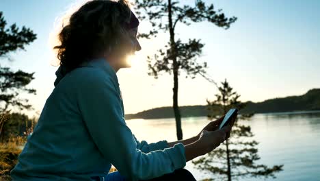 young-woman-uses-smartphone-sitting-on-bank-of-tranquil-lake