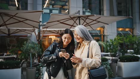 Asian-mature-mum-and-adult-daughter-using-their-smartphones,-looking-at-its-screens-and-laughing-while-standing-near-terrace-of-cafe