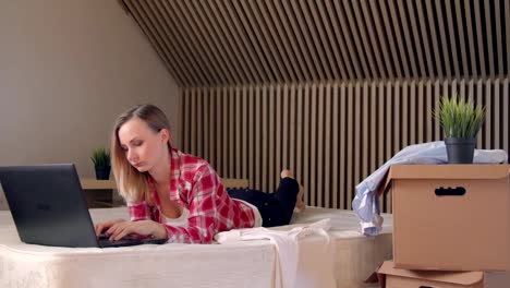 Young-blonde-woman-lying-on-bed-and-working-on-laptop-in-the-morning,-handheld-shot.