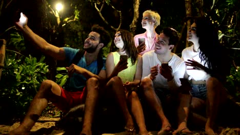 La-gente-toma-selfie-sentado-en-el-banco-en-el-parque-de-la-noche-usando-el-teléfono-celular-inteligente,-amigos-charlando-en-línea-al-aire-libre