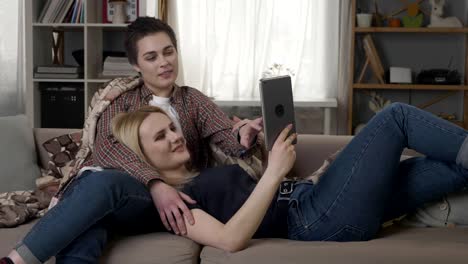 Lesbian-couple-is-resting-on-the-couch,-using-tablet-computer,-scrolling-photos-on-tablet-60-fps