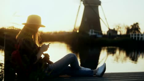 Niña-sentada-con-smartphone-en-el-muelle-del-lago-al-atardecer.-Mujer-con-aire-libre-de-la-aplicación-móvil.-Paisaje-del-molino-de-viento-holandés-tradicional.-4K