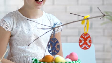 Niña-sonriente-preparando-la-decoración-de-Pascua,-colgantes-huevos-de-juguete-en-las-ramas-de-los-árboles,-Eva