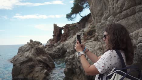 Brunette-woman-is-taking-photos-of-beautiful-rocks-on-seashore-by-mobile-phone