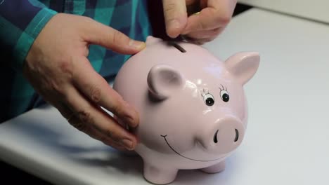 On-the-table-there-is-a-piggy-bank-in-the-form-of-a-pink-pig.-The-man-in-the-hands-of-the-coin,-which-he-puts-in-turn-in-a-piggy-bank.