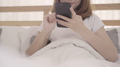 Joven-asiática-con-smartphone-mientras-yacía-en-cama-después-de-despertar-en-la-mañana,-hermosa-chica-japonesa-atractiva-sonriendo-relajarse-en-dormitorio-en-casa.-Disfrutando-de-las-mujeres-de-estilo-de-vida-de-tiempo-en-concepto-de-hogar.