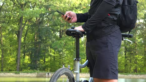 A-cyclist-stands-with-a-bike-on-a-shore-by-a-lake-and-works-on-a-smartphone---closeup