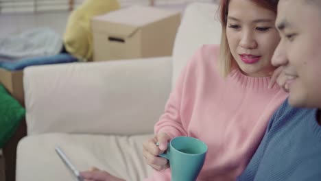 Young-Asian-couple-using-tablet-and-drinking-coffee-in-living-room-at-home,-couple-enjoy-love-moment-while-lying-on-sofa-when-relaxed-at-home.-Enjoying-time-lifestyle-family-at-home-concept.