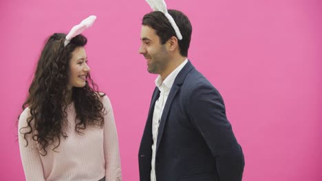 Young-sexy-couple-on-pink-background.-With-hackneyed-ears-on-the-head.-During-this-man-gives-a-soft-toy-with-a-hare-to-his-wife.-Having-kissed-looking-at-the-camera.