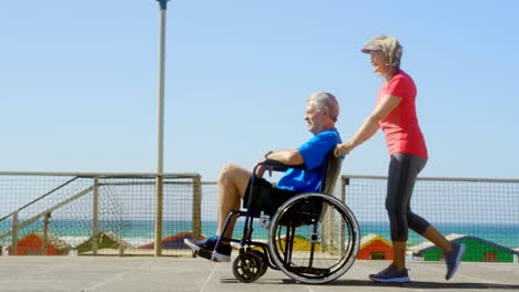 Seitenansicht-der-aktiven-Seniorkaukasschen,-die-ältere-Mann-auf-den-Rollstuhl-am-Strand-4k-drängt