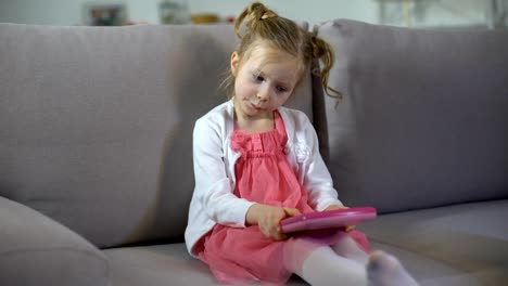 Niña-divertida-sentada-en-el-sofá-y-jugando-en-la-aplicación-educativa-de-la-tableta-de-juguete-rosa