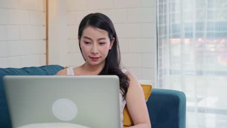 Young-business-freelance-Asian-woman-working-on-laptop-checking-social-media-and-drinking-coffee-while-lying-on-the-sofa-when-relax-in-living-room-at-home.-Lifestyle-women-at-house-concept.