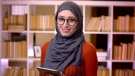 Closeup-shoot-of-young-attractive-muslim-female-student-in-hijab-using-the-tablet-and-looking-at-camera-smiling-standing-indoors-in-the-library