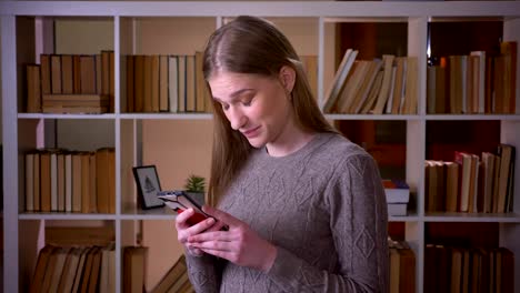 Nahaufnahme-Porträt-von-jungen-attraktiven-Studentin-Browsing-Socail-Medien-am-Telefon-und-Blick-auf-die-Kamera-in-der-College-Bibliothek-in-Innenräumen