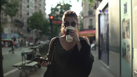 Attractive-caucasian-woman-with-sunglasses,-freckles,-piercings-and-red-hair-scrolling-social-media-on-her-smartphone-and-drinking-coffee-walking-through-the-street,-during-sunny-summer-in-Paris.-4K.