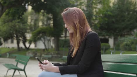 Lächelnde-Geschäftsfrau-mit-Smartphone-Sitzen-auf-der-Bank-im-Park-in-Paris,-SMS-mit-Handy-draußen,-Eingabe-von-SMS-auf-Handy.-Inhalt,-Videos,-Rote-Haare.-Social-Media-im-Freien.