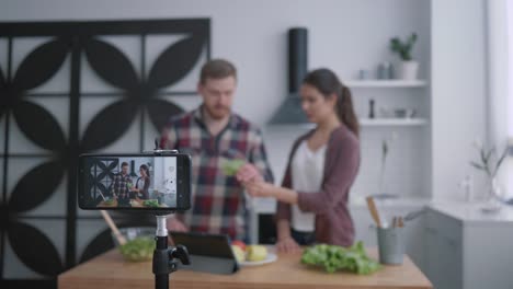 Vegan-Blog,-Vlogger-Kerl-und-Mädchen-bereiten-gesundes-Essen-mit-Gemüse-und-Grüns-in-der-Küche,-während-Kamera-Smartphone-zeichnet-Video-für-Follower-in-sozialen-Netzwerken