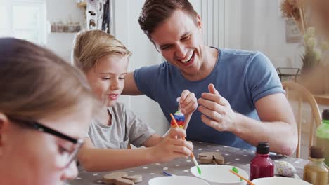 Familie-sitzt-um-Küchentisch-mit-Eltern,-die-Kindern-helfen,-Eier-für-Ostern-zu-malen---in-Zeitlupe-gedreht