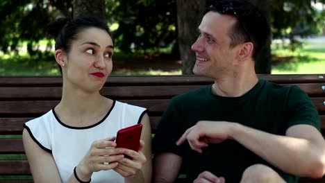 Young-loving-couple,-on-a-sunny-day,-is-actively-discussing-a-new-smartphone-while-sitting-on-a-bench-in-the-park.