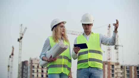 Negocios,-construcción,-industria,-tecnología-y-concepto-de-personas---constructor-sonriente-en-hardhat-con-tableta-PC-ordenador-junto-con-la-mujer-con-dibujos-de-constructores-en-el-sitio-de-construcción.