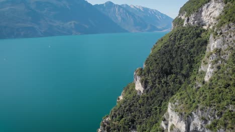 Drohne-fliegt-über-erstaunlichen-Bergsee-mit-türkisfarbenem-Wasser,-entlang-hohen-Berg-bedeckten-Wald-in-sonnigen-Tag