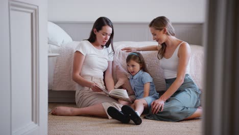 Pareja-femenina-del-mismo-sexo-sentada-en-el-piso-del-dormitorio-libro-de-lectura-con-hija-en-casa-juntos