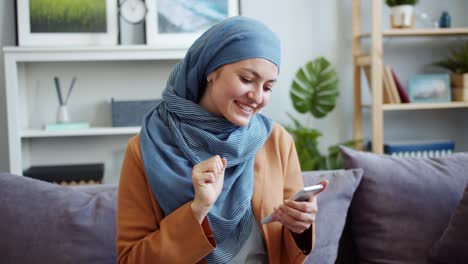 Hermosa-mujer-de-Oriente-Medio-en-hiyab-usando-teléfono-inteligente-en-casa-sonriendo