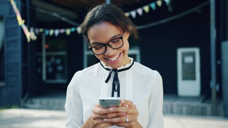 Portrait-of-cheerful-African-American-teen-is-using-smartphone-outdoors-smiling