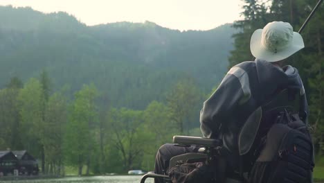 Slow-motion-of-handicapped-fisherman-in-a-electric-wheelchair-fishing-in-beautiful-lake-near-forest-and-mountain-in-the-back,-in-the-sunset