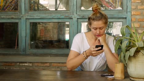 Chica-pensativa-utiliza-un-teléfono-inteligente,-escribe-un-mensaje,-chatea.-Mujer-buscando-información-en-el-teléfono-y-triste.-Sentado-en-el-restaurante-solo.-Hora-del-desayuno.-Concepto-de-tecnología-y-entretenimiento