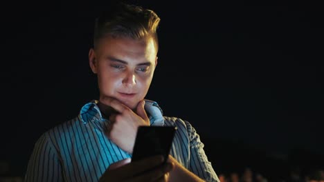 Close-up-portrait-of-young-businessman-using-his-modern-technologies-smartphone-at-night.-Handsome-man-browsing-internet-and-checking-mobile-apps-on-crowded-background.-Illuminated-face-from-phone-light-on-night-background.-Online-apps-and-network