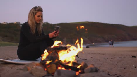 Frau-sitzt-auf-Surfbrett-von-Lagerfeuer-am-Strand-mit-Handy-als-Sonne-setzt-sich-hinter-ihr