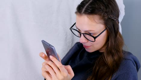 Young-brunette-woman-in-glasses-types-a-message-on-a-mobile-phone-sitting-in-armchair-at-home.