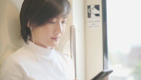 Portrait-of-attractive-smiling-asian-woman-with-earphone-holds-the-handrail-using-smartphone-for-chatting-with-friends-or-browsing-in-public-transport