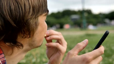 attraktiver-Mann-beißt-Scheibe-Pizza-und-verwendet-Smartphone-Nahaufnahme-im-Park-in-der-Natur