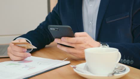 A-businessman-is-sitting-at-a-table-in-a-cafe-and-working.-He-is-entering-his-credit-card-details-on-his-smartphone.-On-the-table-is-a-cup-of-tea-and-documents.-Close-up-shooting.-4K