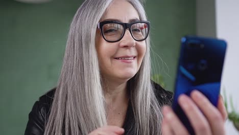 Nahansicht-der-charmanten-glücklichen-Seniorin-in-Brille-mit-langen-grauen-Haaren,-die-Videoanruf-mit-blauem-Telefon