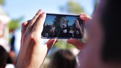 Hand-fingers-hold-phone-making-video-of-wedding-ceremony-screen-close-up-slow-motion.-Smart-technologies-live-event-social-media-networks-guest-using-device-to-record-friends-couple-getting-married