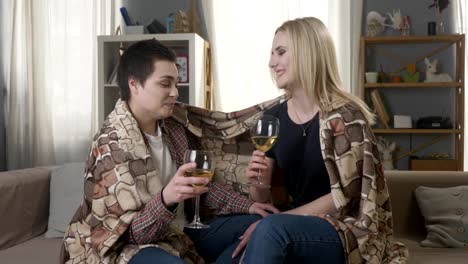 Dos-chicas-jóvenes-y-hermosas-están-sentados-en-el-sofá-envoltura-con-manta-plaid,-bebiendo-vino-y-riendo-60-fps