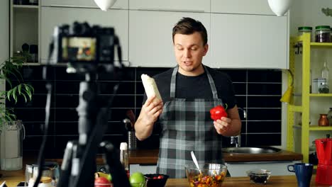 Cheerful-attractive-man-recording-video-food-vlog-about-healthy-cooking-on-digital-camera-in-the-kitchen-at-home.-Vlogging-and-social-media-concept