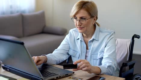 Happy-adult-woman-in-wheelchair-entering-card-number-on-laptop,-shopping-online