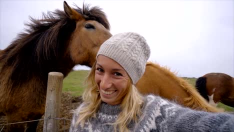 Blond-hair-girl-in-Iceland-taking-selfie-portrait-with-Icelandic-horse-in-green-meadow.-Shot-in-Springtime,-overcast-sky,-woman-wearing-Icelandic-grey-wool-pullover.-People-travel-animal-affection-concept--Slow-motion