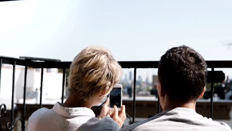 Happy-romantic-couple-sitting-at-a-small-sunny-balcony,-taking-smartphone-photos-of-amazing-Manhattan-view-in-New-York