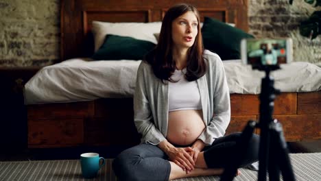 Pretty-pregnant-woman-famous-blogger-is-recording-video-for-followers-looking-at-smartphone-camera-and-talking-sitting-on-floor-in-bedroom-and-smiling.